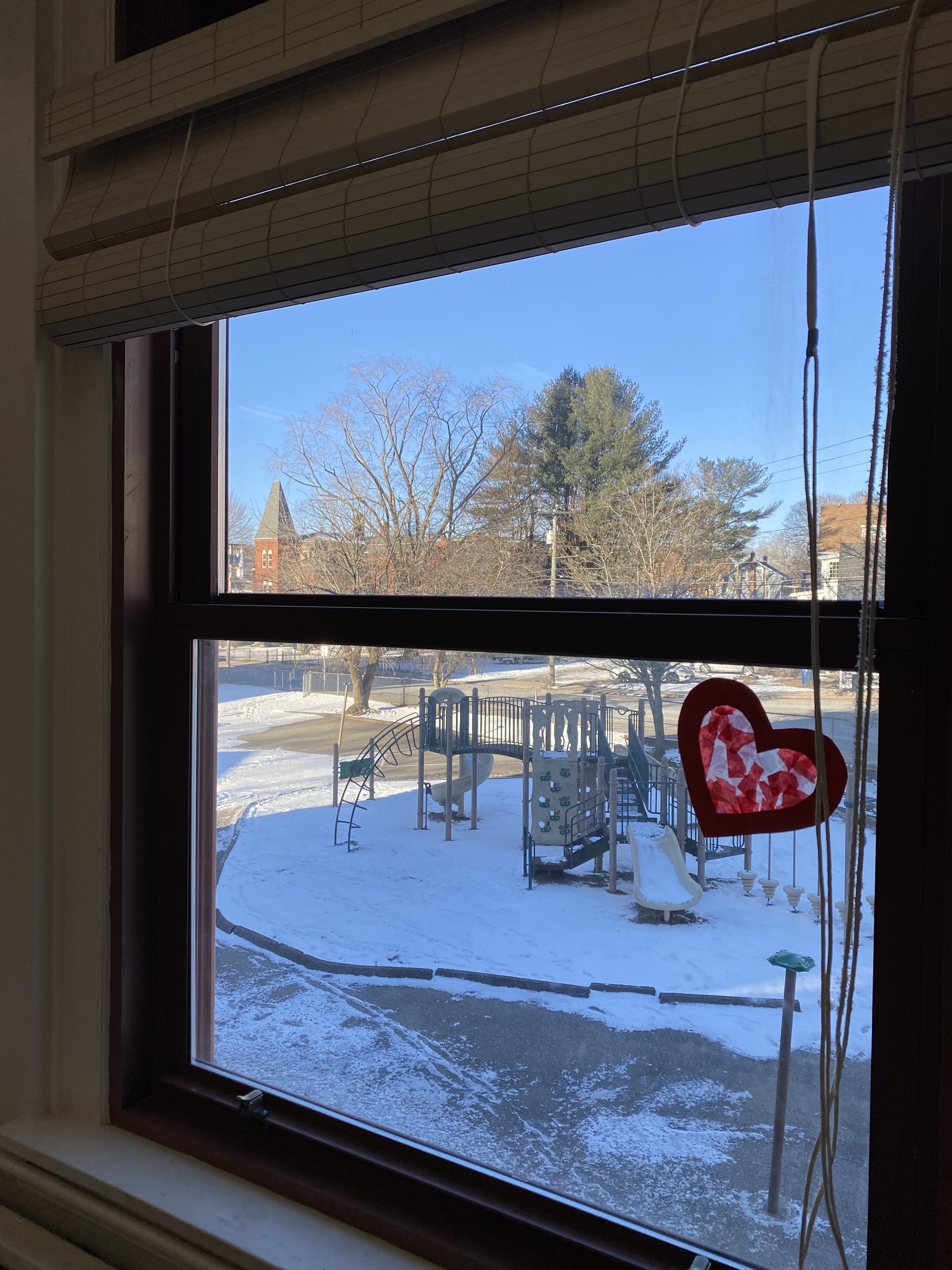  This is an image of the view out of a window of a deserted snowy playground from an upper story window. There is a heart decal on the bottom window frame made of tissue paper. 